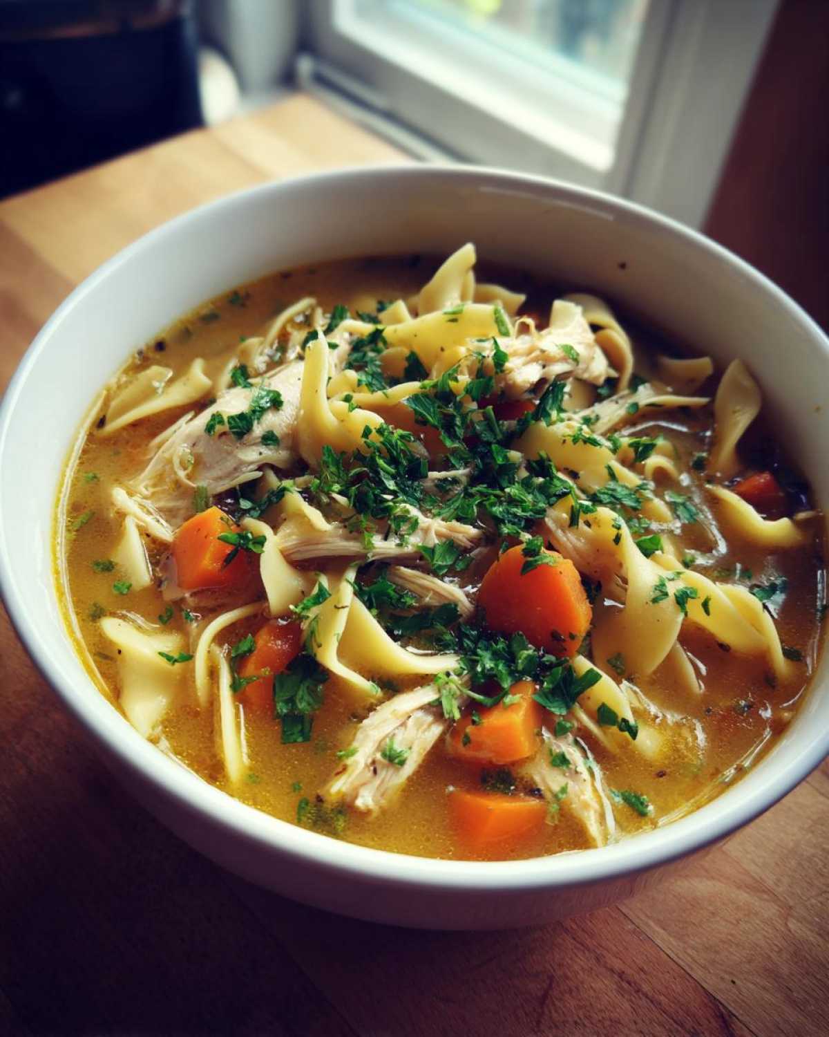 Close-up of a bowl of rich slow cooker chicken noodle soup, featuring egg noodles, shredded chicken, carrots, and fresh parsley.