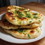 A stack of three soft, freshly made naan bread pieces brushed with melted butter and topped with chopped green herbs.