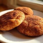 Close-up of three soft snickerdoodle cookies heavily coated in cinnamon sugar, resting on a white plate near a window.