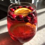 Close-up of a glass filled with sparkling red non-alcoholic christmas punch garnished with fresh cranberries and orange slices.