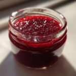 Close-up of a small glass jar filled with bright, glistening homemade raspberry jam, catching the sunlight.