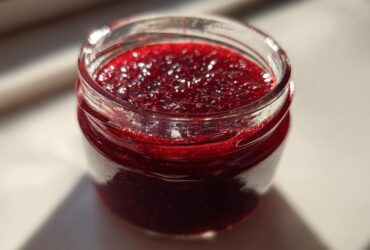 Close-up of a small glass jar filled with bright, glistening homemade raspberry jam, catching the sunlight.