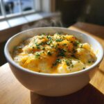 A steaming white bowl filled with chunky easy crockpot potato soup, topped generously with melted cheddar cheese and fresh chives.