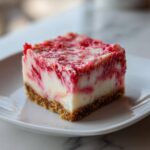 A close-up of a square slice of strawberry crunch cheesecake with a graham cracker crust.