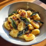 A bowl of golden, pan-seared sweet potato gnocchi topped with crispy sage leaves and grated cheese.