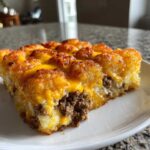 A close-up slice of baked tater tot casserole topped with melted cheddar cheese and ground beef filling.