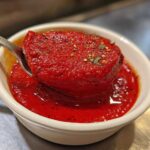 Close-up of thick, vibrant red pizza sauce being scooped with a spoon from a small white bowl.