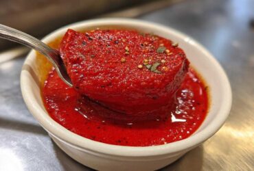 Close-up of thick, vibrant red pizza sauce being scooped with a spoon from a small white bowl.