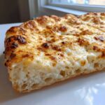 Close-up cross-section of a thick, fluffy crust topped with creamy, bubbly white sauce and browned cheese on a white pizza.