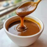 Close-up of thick, rich homemade brown gravy dripping from a golden spoon back into a white bowl.