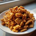 A close-up of a mound of glistening, golden-brown toffee chex mix recipe pieces on a white plate.