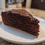 Close-up of a dense, dark slice of moist chocolate cake resting on a white plate.