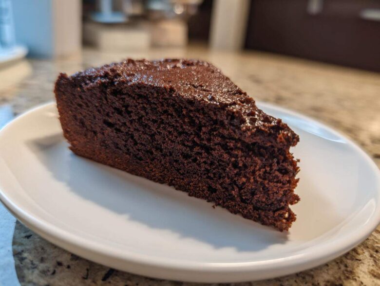 Close-up of a dense, dark slice of moist chocolate cake resting on a white plate.