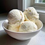 Four scoops of creamy vanilla protein ice cream served in a white bowl next to a sunlit window.