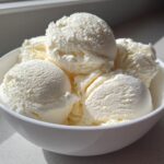 Several scoops of creamy vanilla protein ice cream filling a white bowl, brightly lit by sunlight.