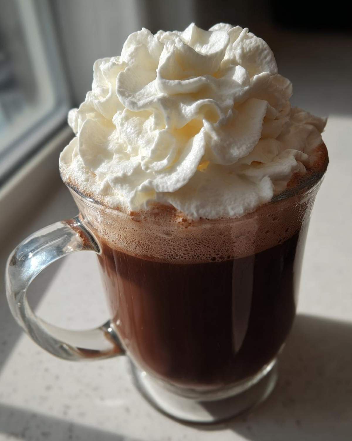 A clear glass mug filled with rich, dark whipped hot chocolate topped with a generous swirl of whipped cream.