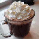 A clear glass mug filled with rich, dark whipped hot chocolate topped generously with fluffy whipped cream.