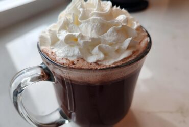A clear glass mug filled with rich, dark whipped hot chocolate topped generously with fluffy whipped cream.