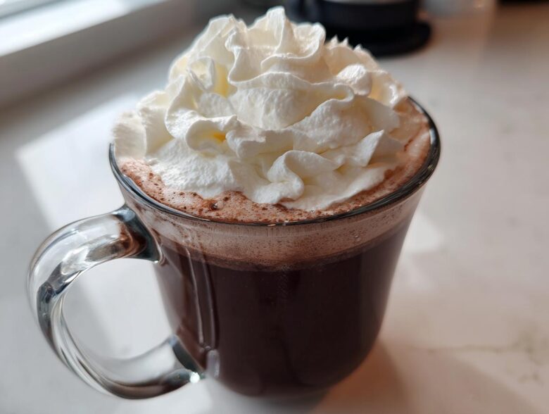A clear glass mug filled with rich, dark whipped hot chocolate topped generously with fluffy whipped cream.