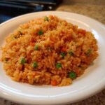 A close-up of a serving of vibrant orange spanish rice mixed with bright green peas and diced carrots on a white plate.