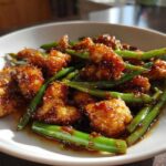 Close-up of crispy, glazed chicken pieces mixed with bright green beans in a savory sauce, featuring chicken and green beans.