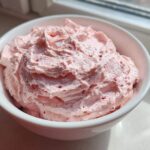 A close-up of fluffy, light pink strawberry cheesecake dip swirled high in a white bowl, sitting near a window.