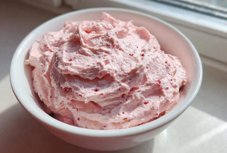 A close-up of fluffy, light pink strawberry cheesecake dip swirled high in a white bowl, sitting near a window.