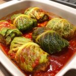 Close-up of six homemade cabbage rolls simmering in a rich, bright red tomato sauce in a white baking dish.