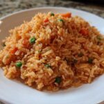 A close-up of fluffy, orange-red spanish rice mixed with bright green peas and diced carrots on a white plate.