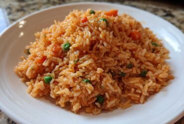 A close-up of fluffy, orange-red spanish rice mixed with bright green peas and diced carrots on a white plate.