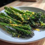 A white plate featuring bright green, roasted broccolini topped with visible lemon zest and coarse salt.