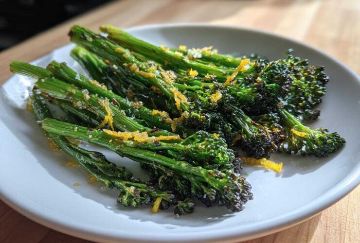 A white plate featuring bright green, roasted broccolini topped with visible lemon zest and coarse salt.