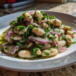 A close-up of a vibrant white bean salad mixed with red onion slices and fresh green herbs, sitting in dressing on a white plate.