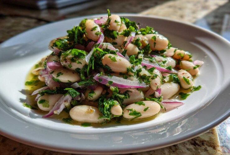 A close-up of a vibrant white bean salad mixed with red onion slices and fresh green herbs, sitting in dressing on a white plate.