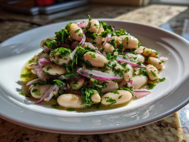 A close-up of a vibrant white bean salad mixed with red onion slices and fresh green herbs, sitting in dressing on a white plate.