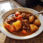 A close-up of a white bowl filled with rich, dark brown pork stew chunks, potatoes, and carrots.