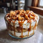 Close-up of a layered caramel apple trifle featuring whipped cream, spiced apples, and heavy caramel drizzle.