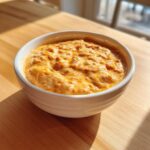 A close-up of warm, creamy, orange beer cheese dip served in a white bowl on a sunlit wooden table.