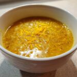 A close-up of a vibrant yellow chickpea soup garnished with orange zest in a white bowl.