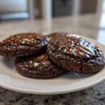 Close-up of three rich, dark chocolate brownie cookies with glossy, cracked tops on a white plate.