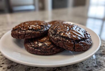 Close-up of three rich, dark chocolate brownie cookies with glossy, cracked tops on a white plate.