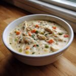 A close-up of a hearty bowl of homemade cream of chicken soup, filled with shredded chicken, carrots, and peas.