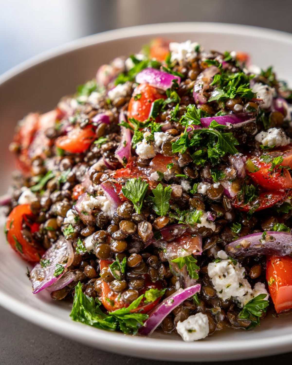 A close-up, appetizing shot of a vibrant lentil salad featuring brown lentils, chopped tomatoes, red onion, parsley, and crumbled feta cheese.