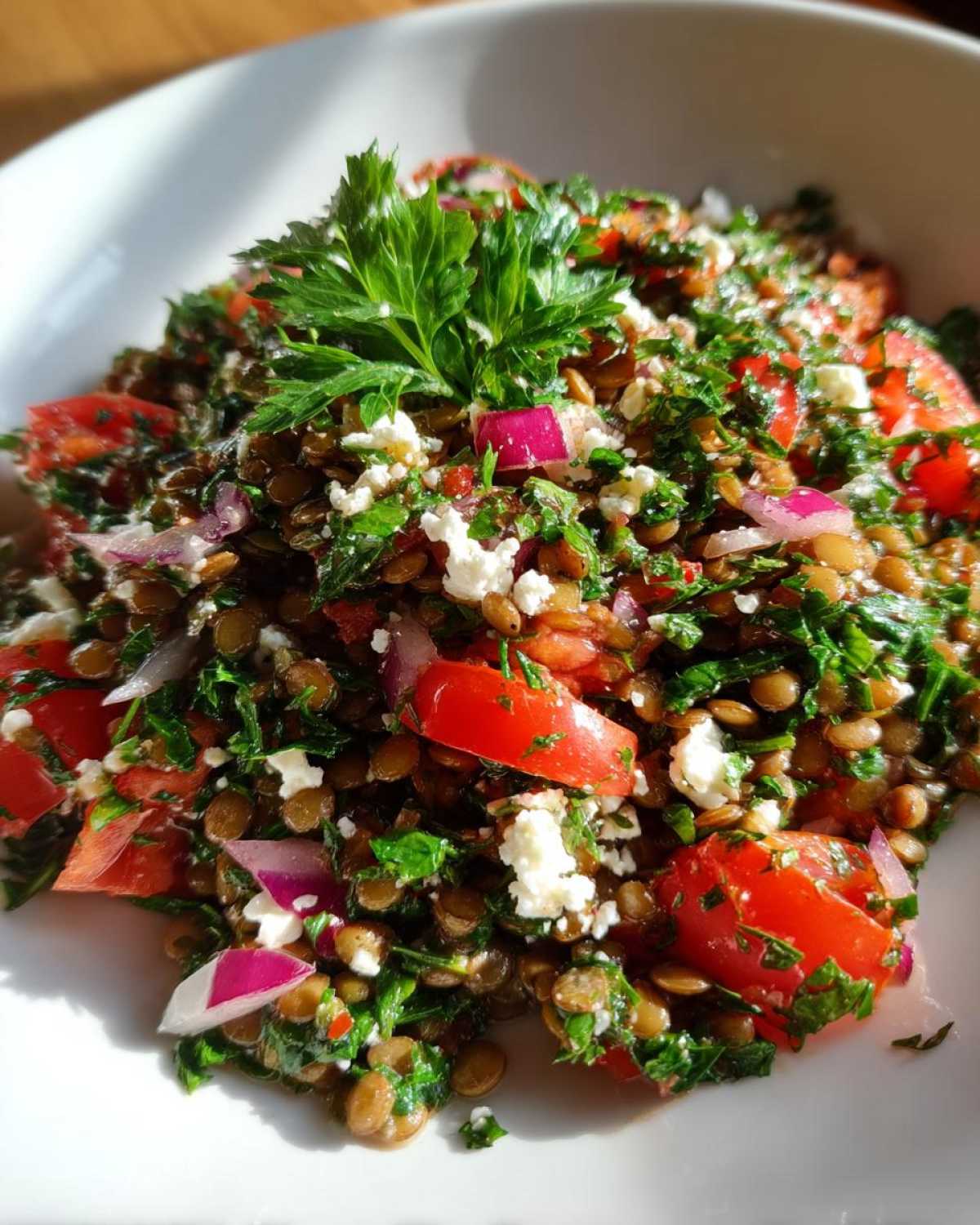Close-up view of a vibrant lentil salad mixed with chopped tomatoes, red onion, feta cheese, and parsley garnish.