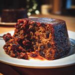 Close-up of a rich, dark slice of plum pudding with visible dried fruit, served on a white plate.