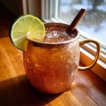 A frosty copper mug filled with an apple cider moscow mule, garnished with a lime wedge and cinnamon sticks.
