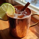 A frosty copper mug filled with an apple cider moscow mule, garnished with a lime wheel and cinnamon stick.