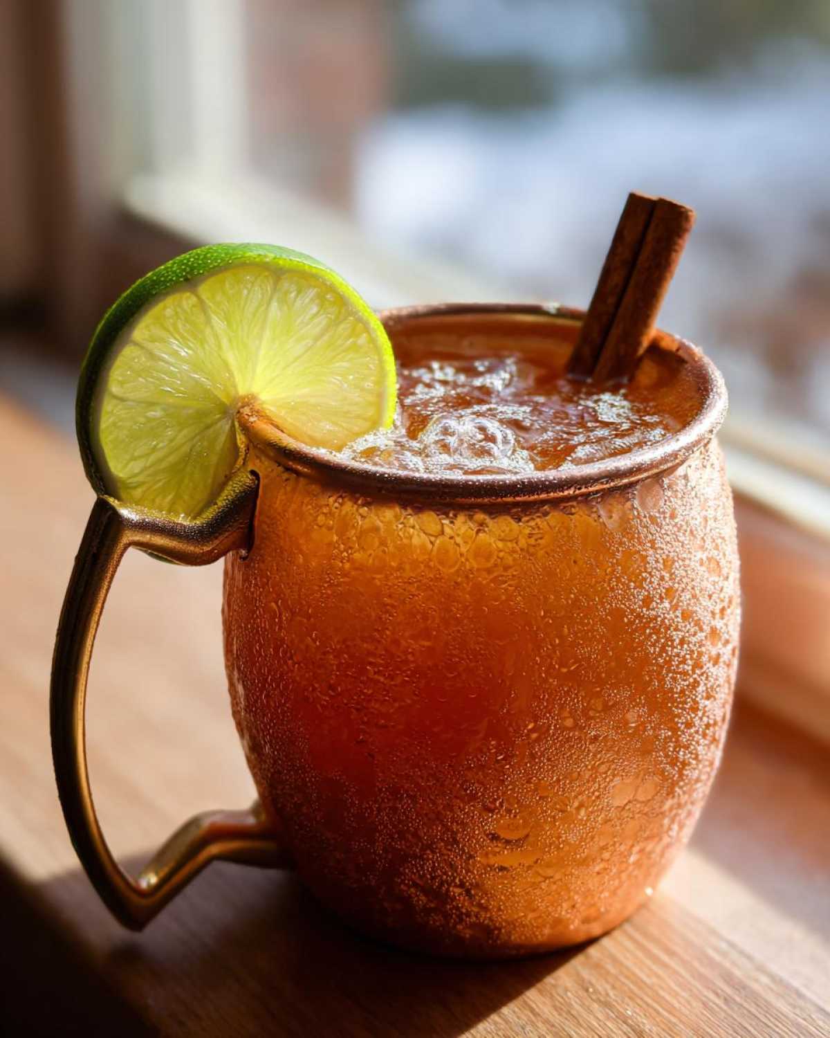 A close-up of a frosty copper mug filled with an apple cider Moscow mule, garnished with a lime wheel and cinnamon stick.