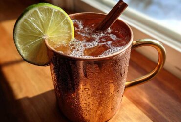 A frosty copper mug filled with an apple cider moscow mule, garnished with a lime wheel and cinnamon stick.