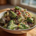 A close-up of a creamy broccoli salad featuring fresh broccoli florets, bacon bits, dried cranberries, and slivered almonds.